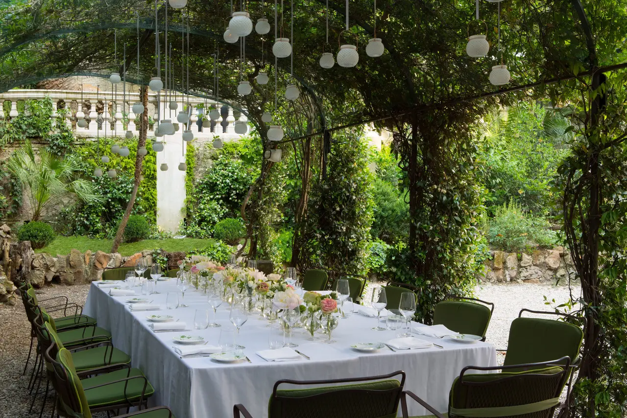 A long table set under the pergola in the Secret Garden: flowers, glasses, green chairs and white glass lamps hanging among the climbing plants; behind, a lawn and a balustrade
