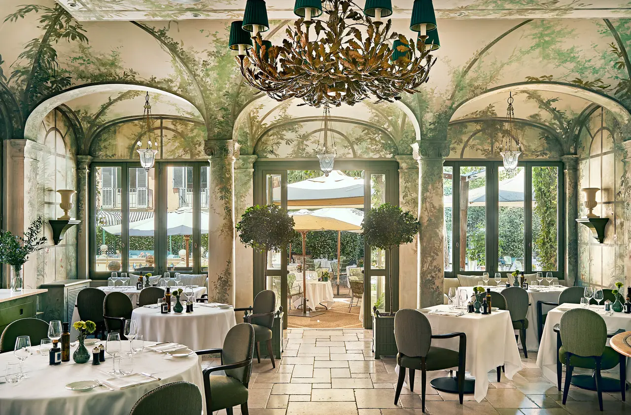 Le Jardin’s dining room: walls and vaults painted with climbing plants, a gilded leaf chandelier with green shades, round tables with white cloths and sage-green chairs; the French windows open onto the terrace and its parasols
