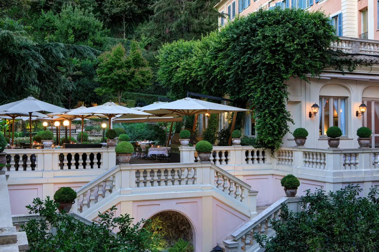 Le Jardin’s terrace on a summer evening: tables with white cloths and lit candles under the parasols, globe lamps and box balls in terracotta urns on the white balustrade; below, the double staircase