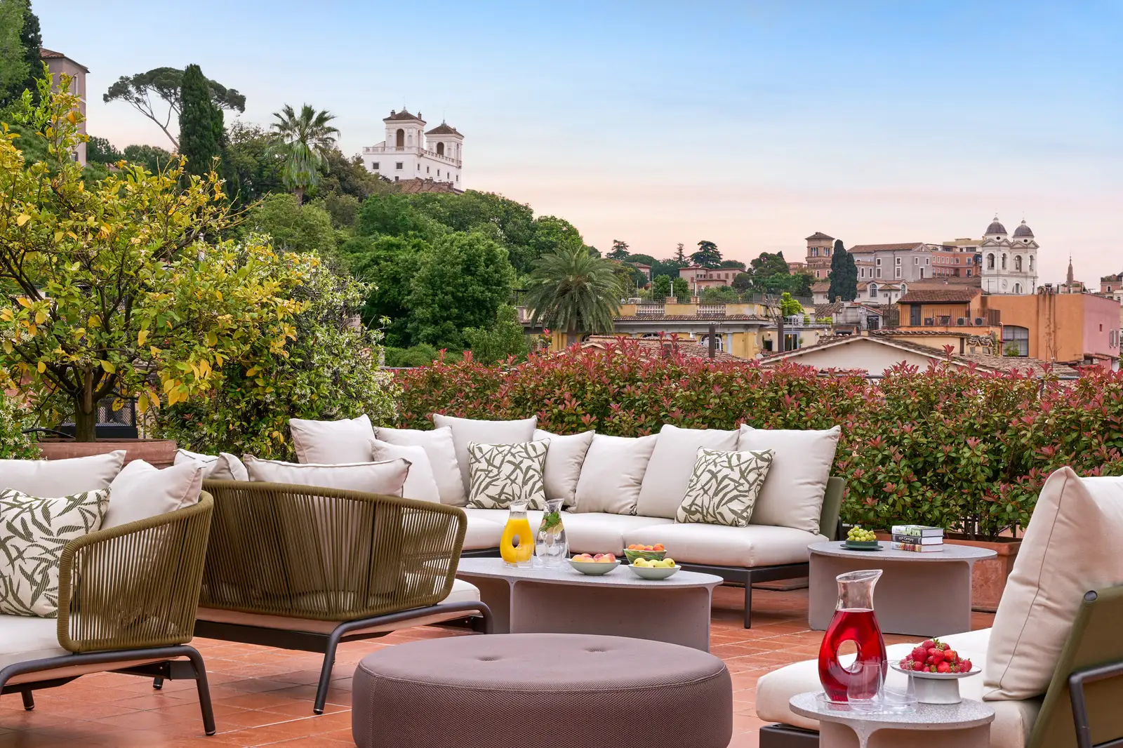 The terrace of the Nijinsky Suite: white sofas and rope armchairs on terracotta tiles, low tables with fruit and a carafe, a red-leaved hedge and a potted citrus tree; beyond the rooftops, the trees of the Pincio, Villa Medici and the twin bell towers of Trinità dei Monti