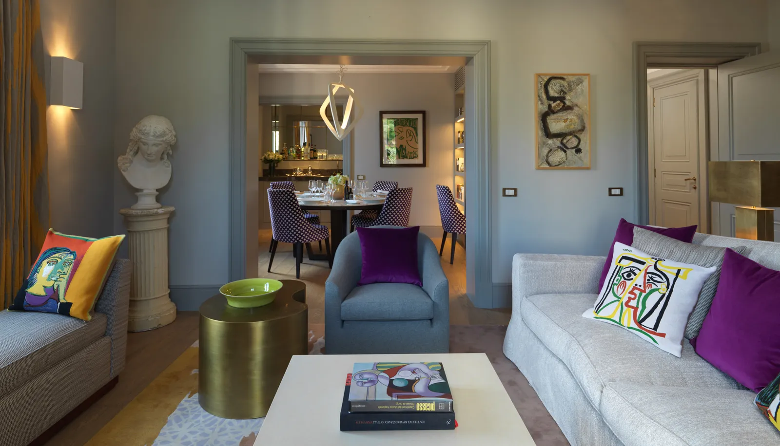 The sitting room of the Picasso Suite: grey-blue walls, a classical bust on a column, sofas with cushions printed with Cubist-style faces and purple velvet cushions; through the doorway, the dining room with its round table