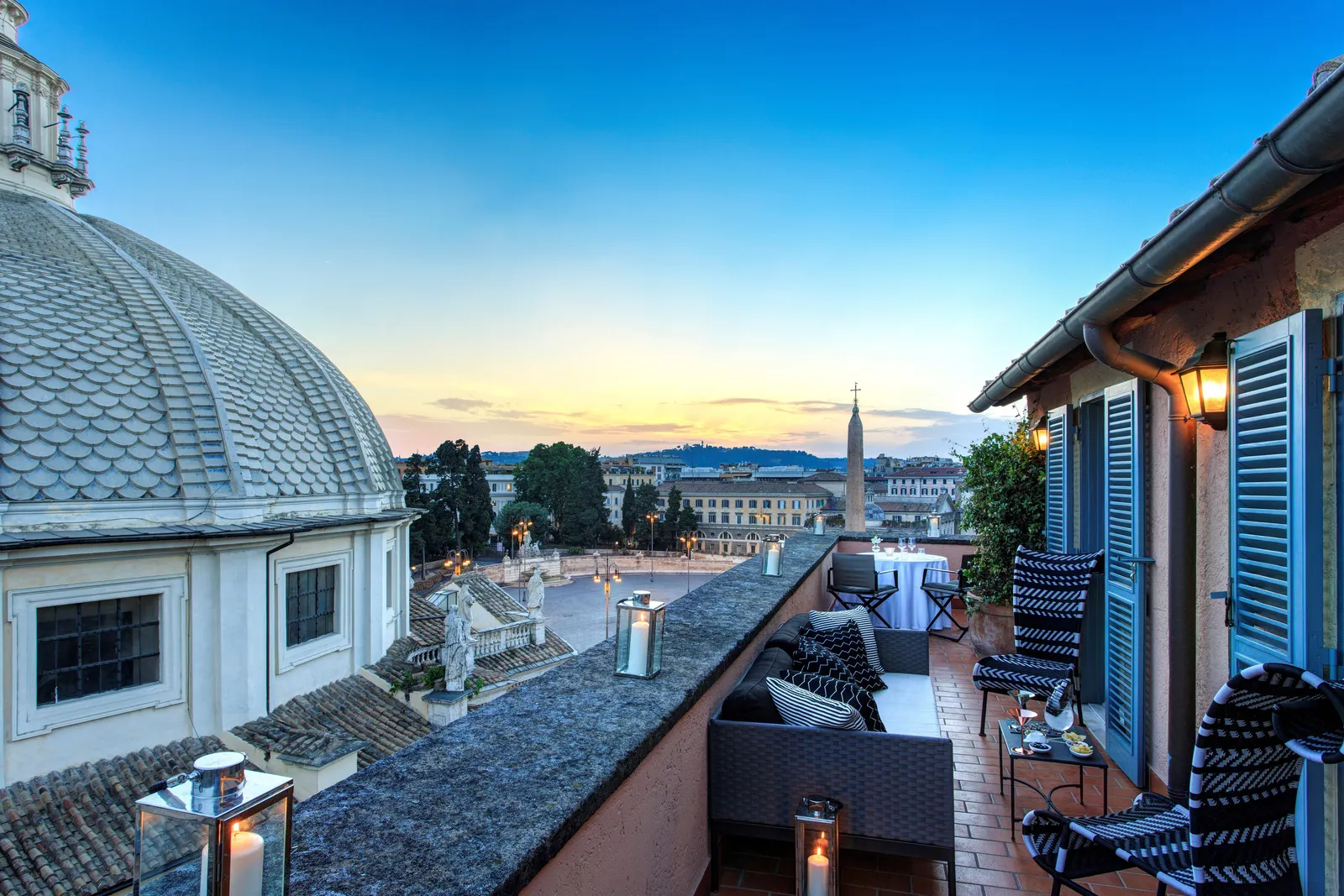 The Popolo Suite’s terrace at sunset: woven sofas and armchairs with black-and-white cushions, lit lanterns on the parapet and blue shutters; on the left the dome of one of the twin churches, and beyond, the obelisk of Piazza del Popolo and the rooftops out to the hills