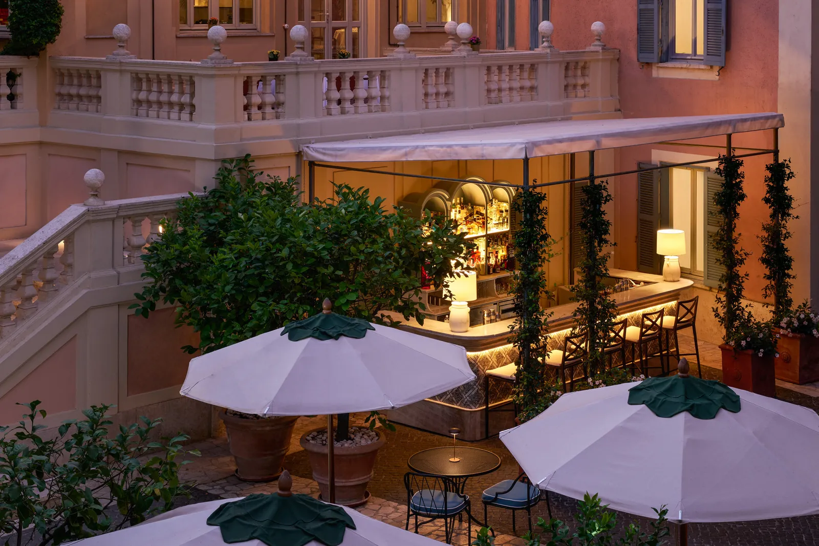 The Stravinskij Bar from above at dusk: 2 white parasols with green tops, the lit counter under its awning, potted trees and, above, the balustraded terrace of the pink building