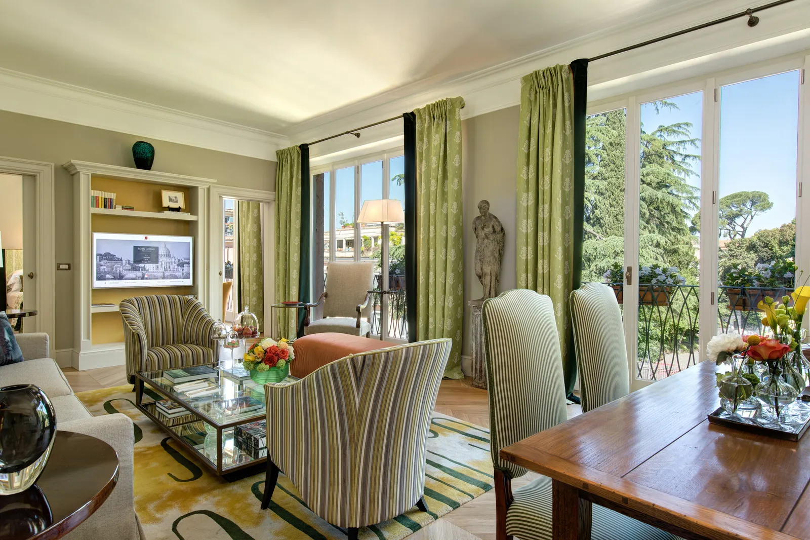 The sitting room of the Valadier Suite: French windows with green curtains open onto a flower-lined balcony and umbrella pines, striped armchairs around a glass coffee table, a wooden dining table and a classical statue by the window