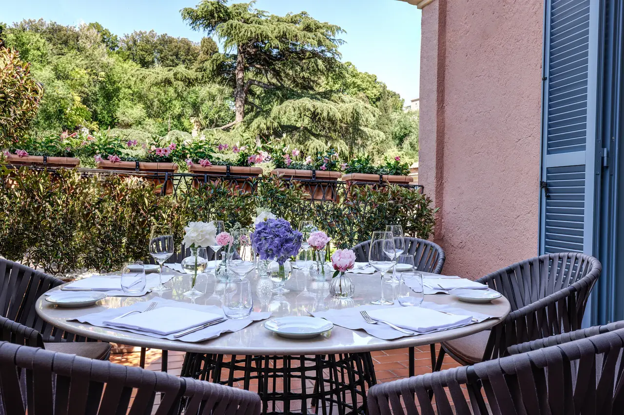 The terrace of the Vaselli Suite: a laid round table with hydrangeas and peonies at its centre, dark woven chairs, planters of pink flowers along the railing and the garden’s trees beyond