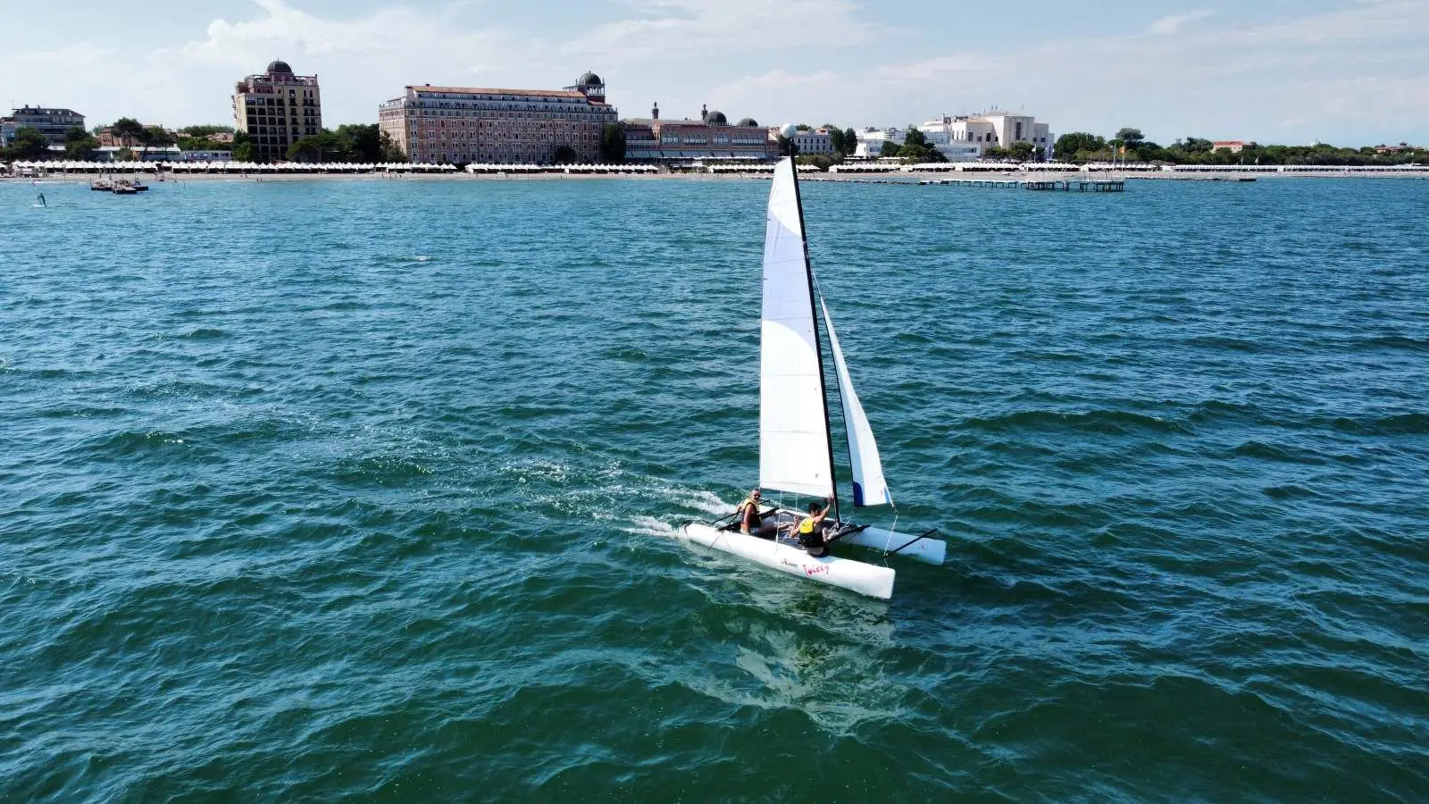 A small sailing catamaran on the green sea, with the beach, the cabanas and the Excelsior’s façade on the shore.