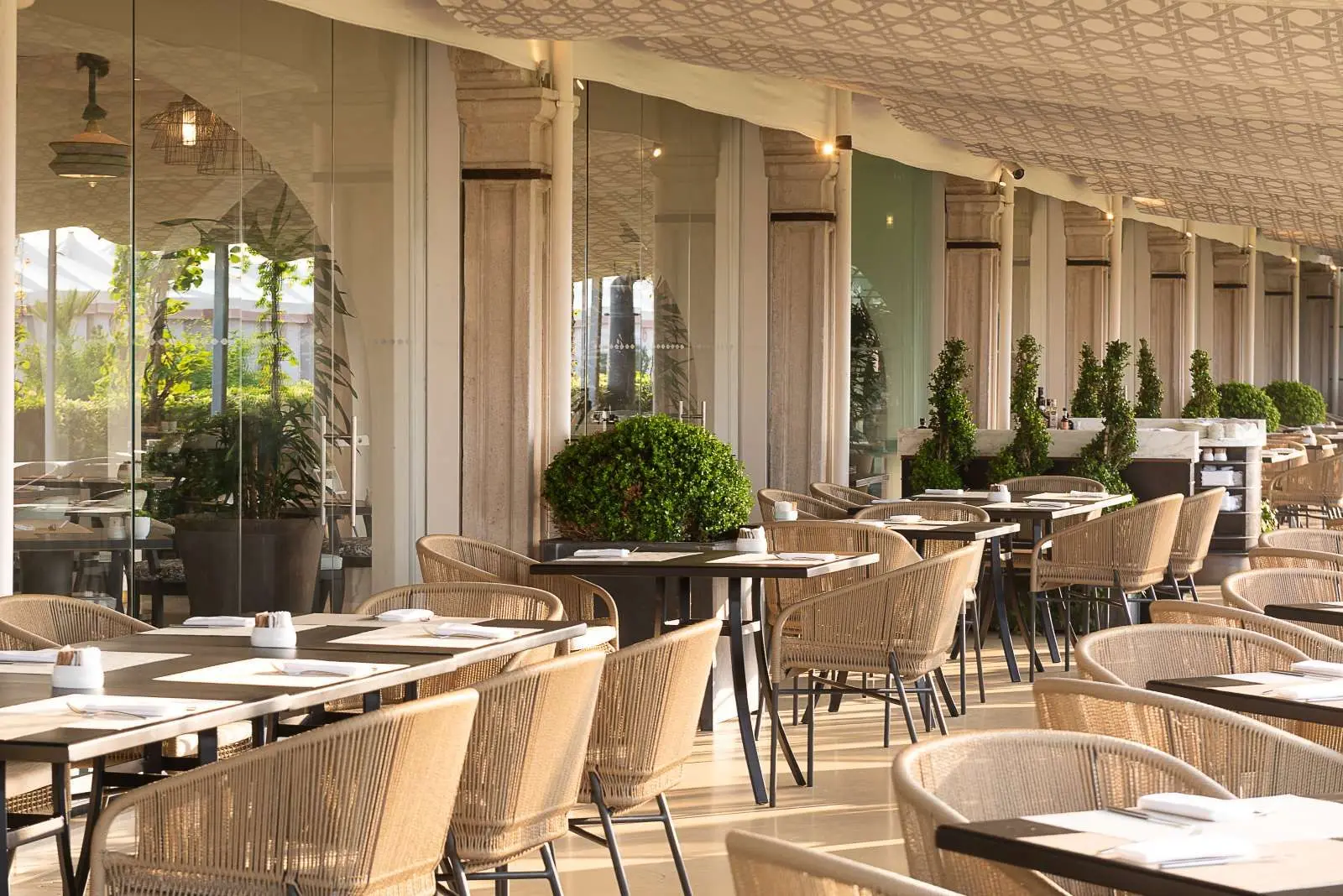 Elimar’s veranda: laid tables and woven-rope armchairs along the glass wall, under a geometric-patterned ceiling.