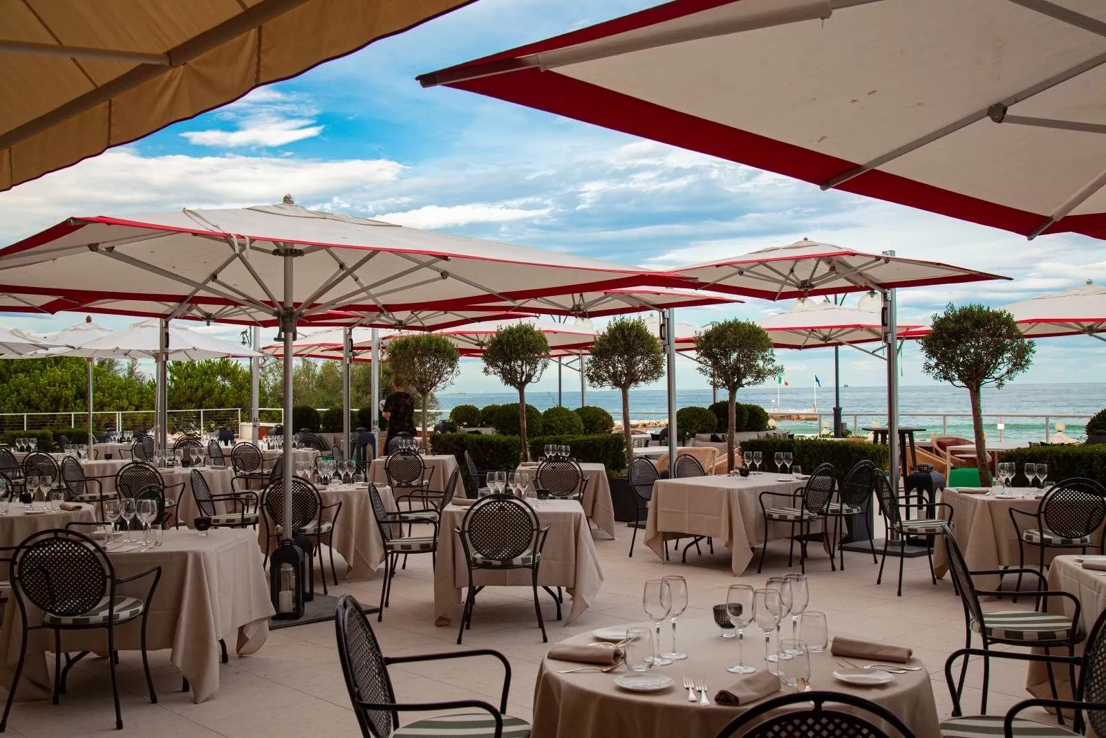 The restaurant terrace by the sea: laid tables under red-trimmed umbrellas, potted trees and the Adriatic beyond the hedge.