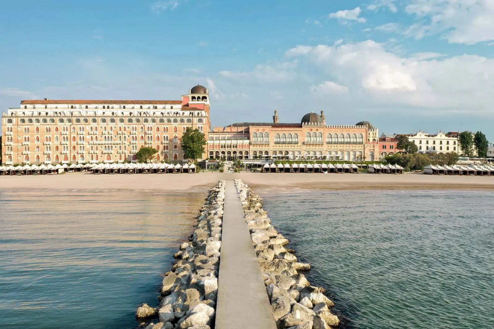 The stone pier leading from the sea to the beach, with the cabanas in a row and the Excelsior’s façade under a cloudy sky.