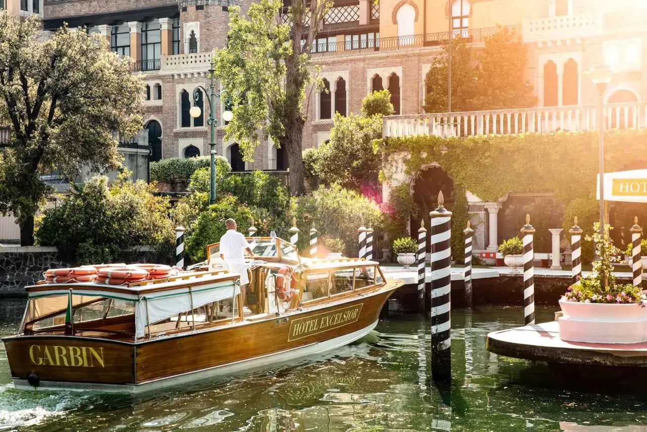 A wooden launch marked Hotel Excelsior pulls up to the canal dock, between black-and-white mooring poles and the garden, in sunset light.