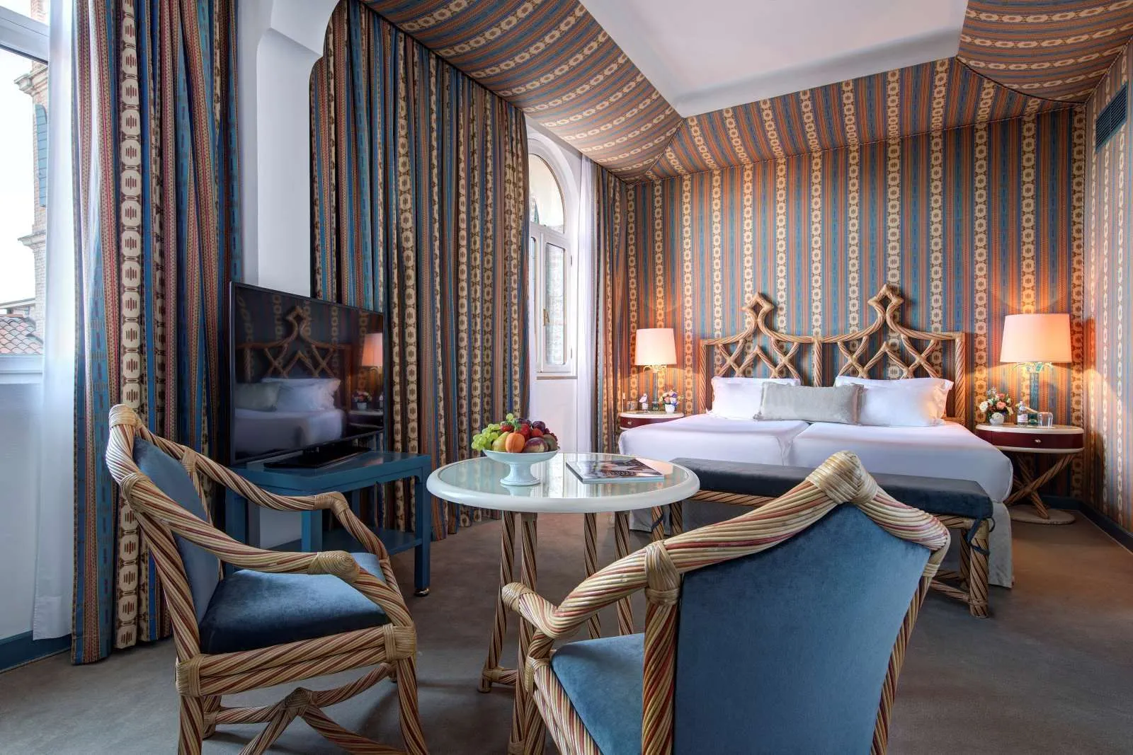 A room with twin beds pushed together under a striped fabric canopy, woven wooden headboards, a round table and 2 rattan armchairs.