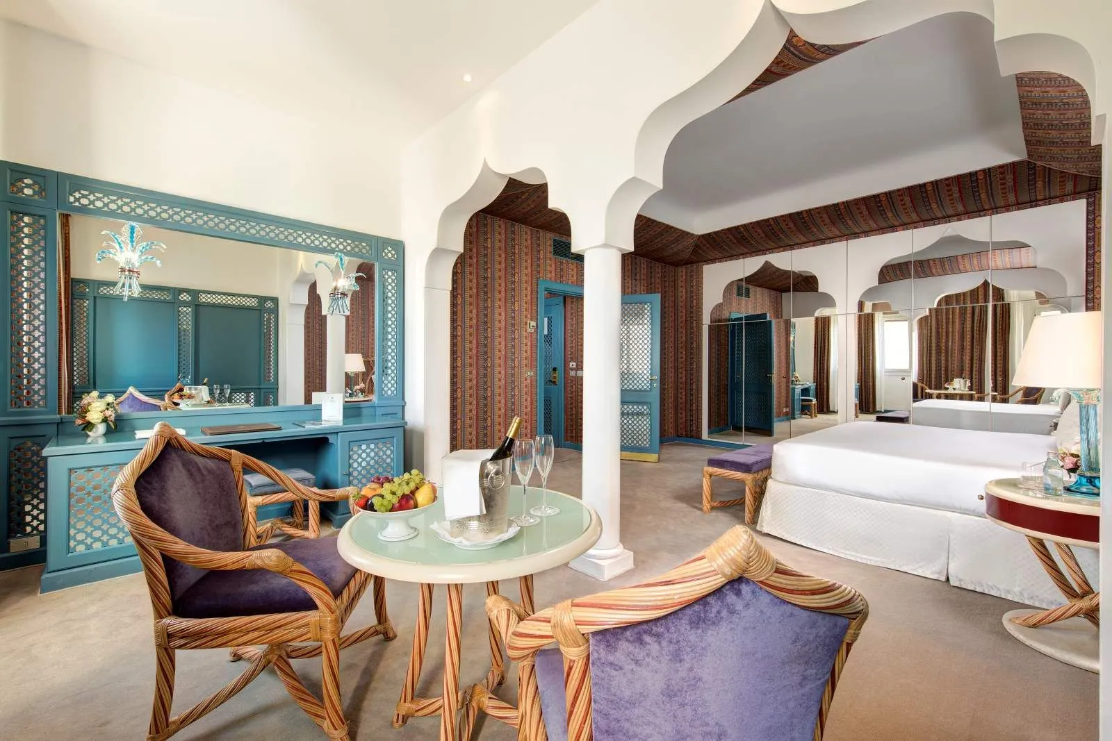 A large room divided by Moorish arches: a table with sparkling wine and fruit, purple armchairs, a blue desk and the bed beyond.