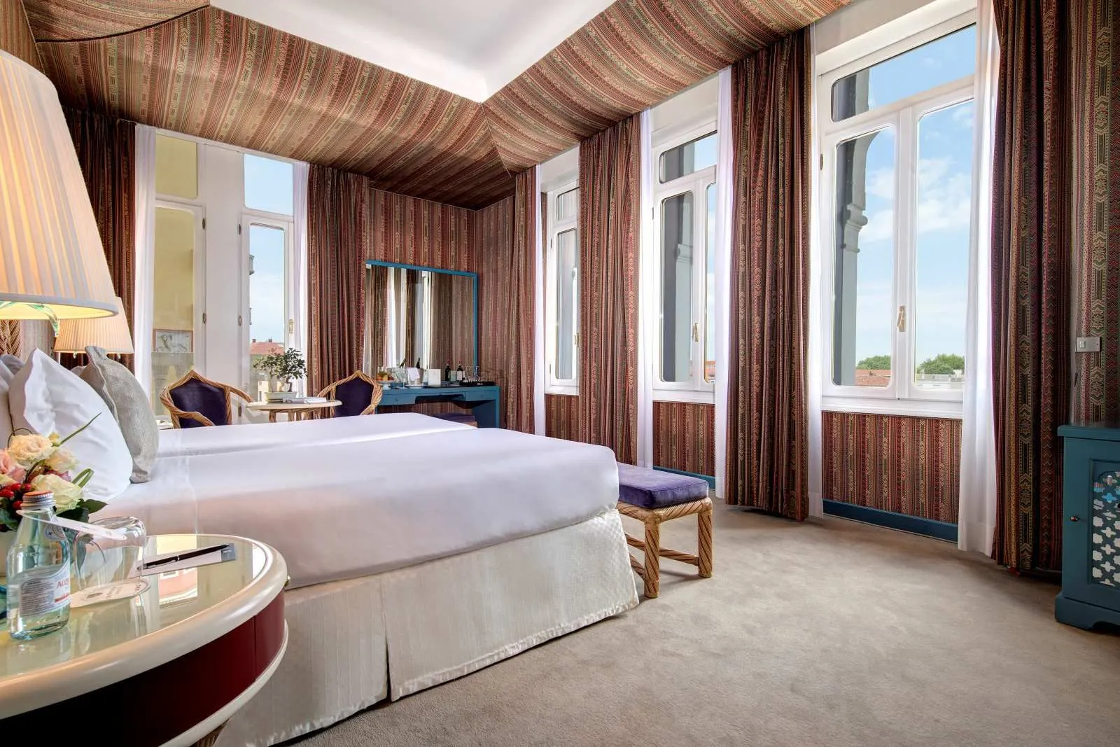 A room with a king bed, 3 tall windows onto sky and trees, and walls and ceiling lined in striped fabric.