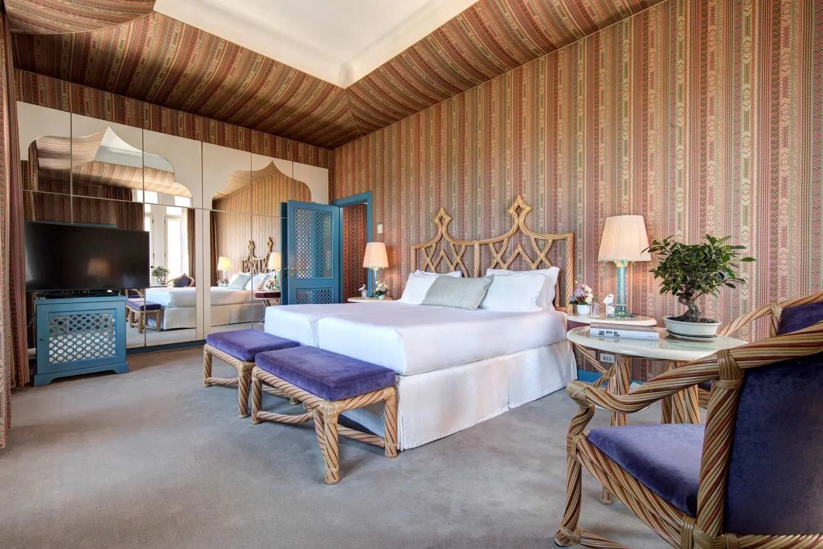 A room with a king bed and woven wooden headboard, striped walls, a mirrored wall and 2 purple armchairs.