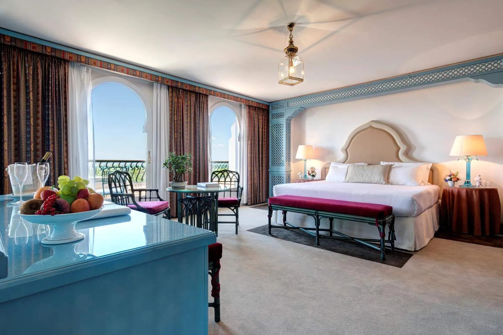 A room with a king bed, a red bench at its foot, a blue desk with fruit and 2 arched French windows open onto the balcony.