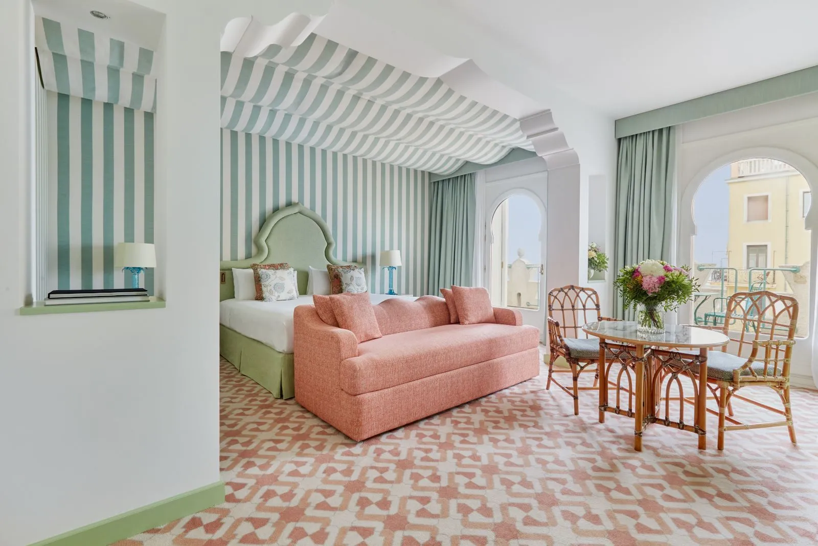 A green-striped room with the bed, a pink sofa, a round table with flowers and arched windows over the rooftops.