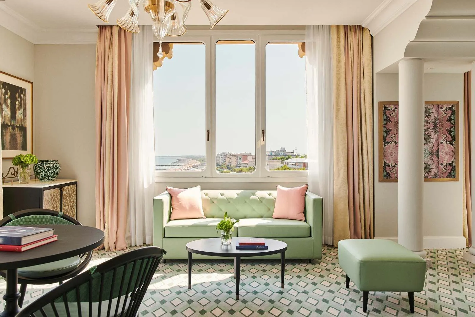 The Junior Suite’s sitting room: a green sofa with pink cushions, a round table, a glass chandelier and 3 windows over the shore.