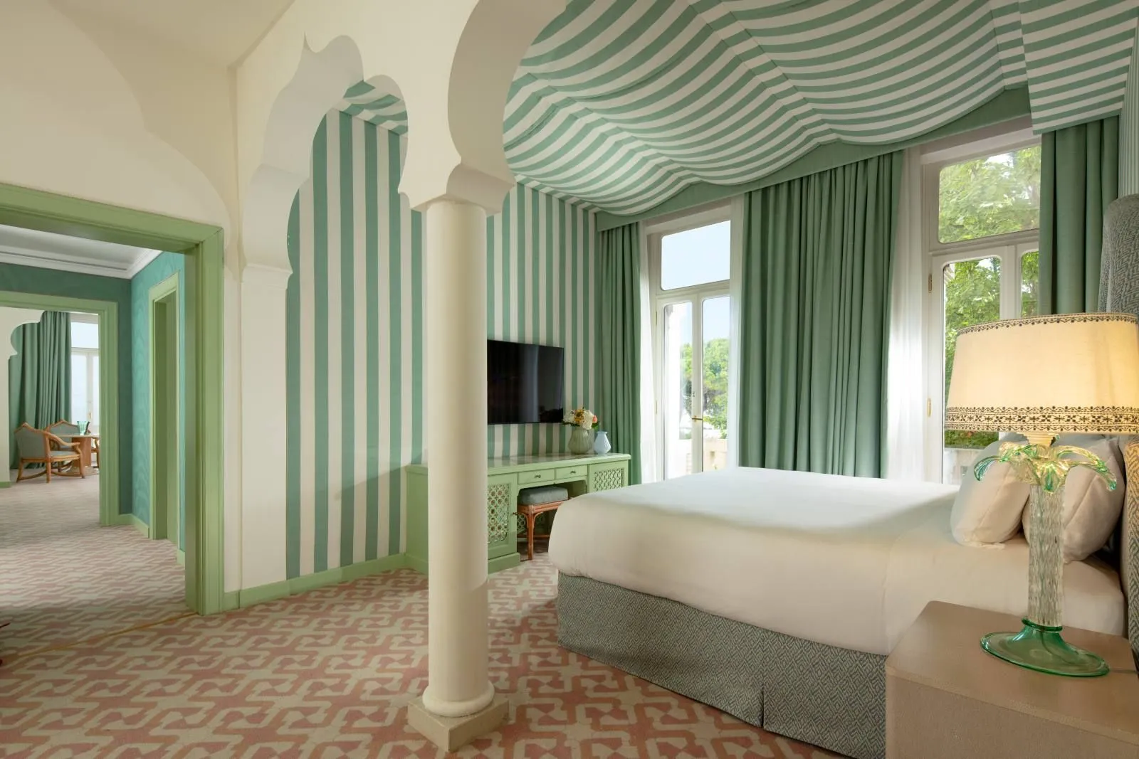 A room with a green-striped tented ceiling, a column and arches, the bed and windows onto trees; through the door, the second room.