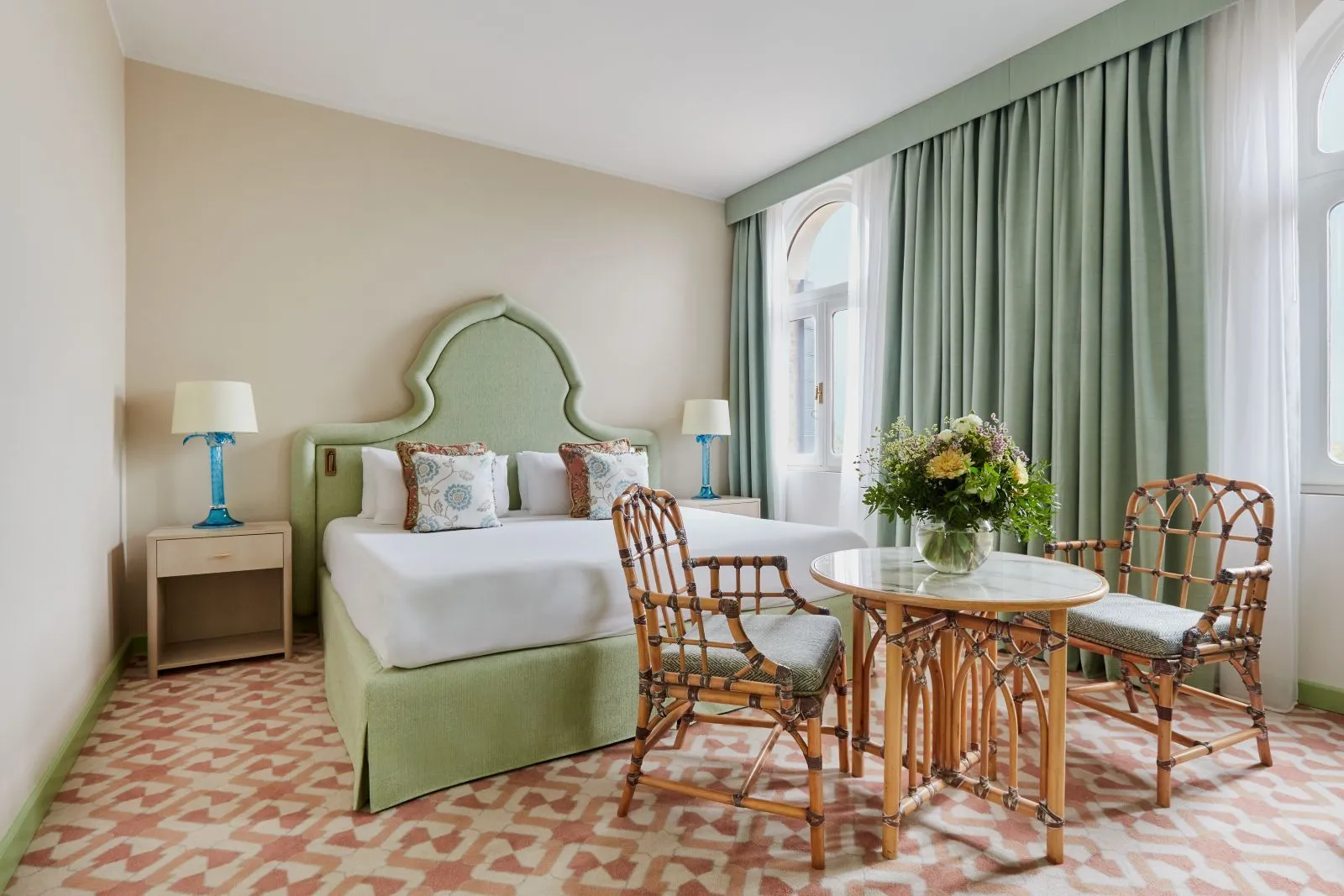 A bright room with a mint-green headboard, green curtains, a pink-patterned carpet and a small table with 2 rattan chairs.