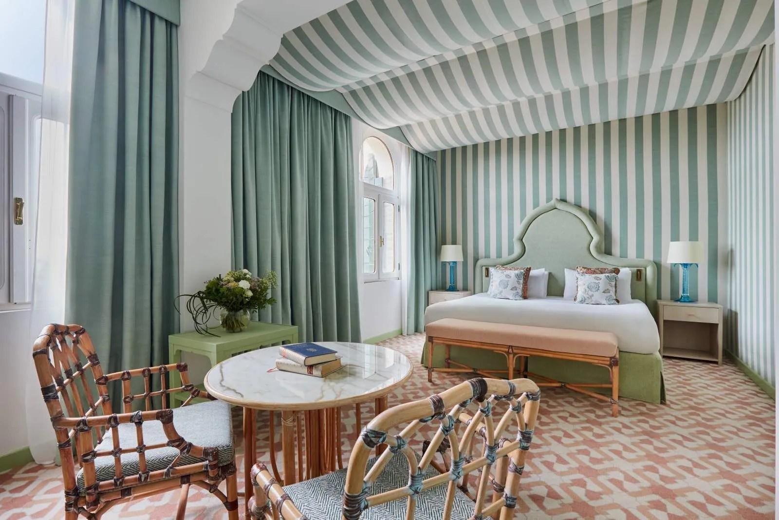 A room with a green-and-white striped tented ceiling, a green headboard, a pink bench and a marble table with rattan armchairs.
