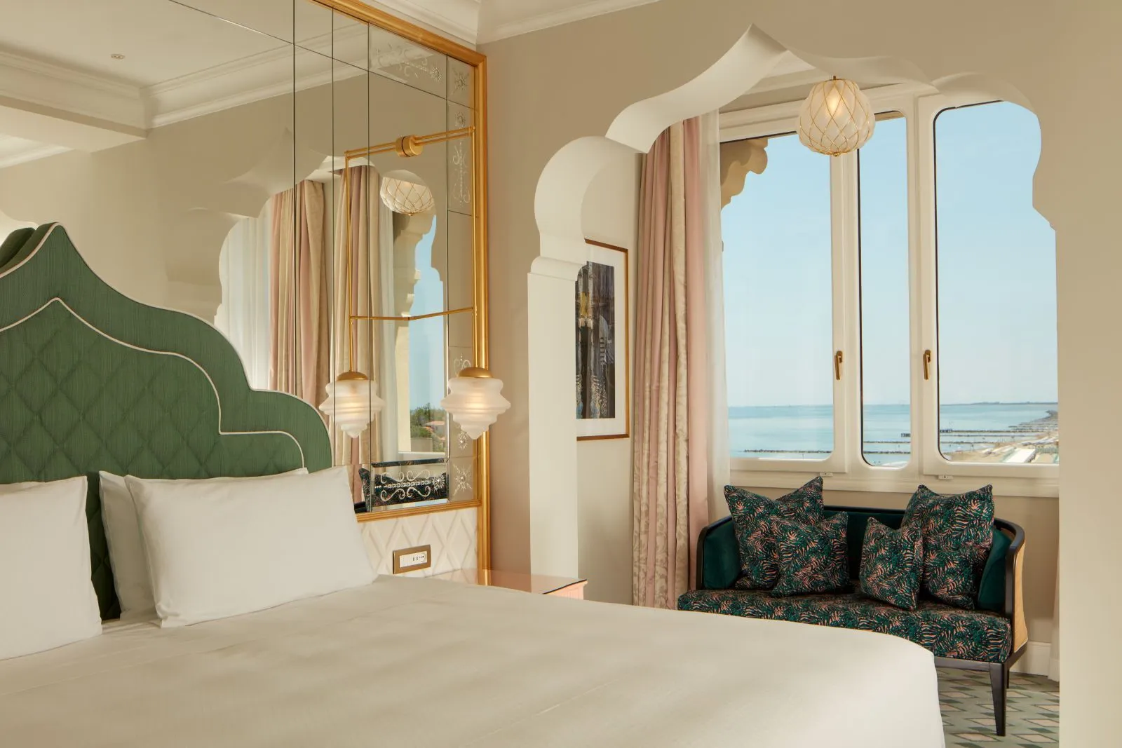 The bed with a quilted green headboard, a gilt mirror and a small sofa under arched windows looking at the sea.