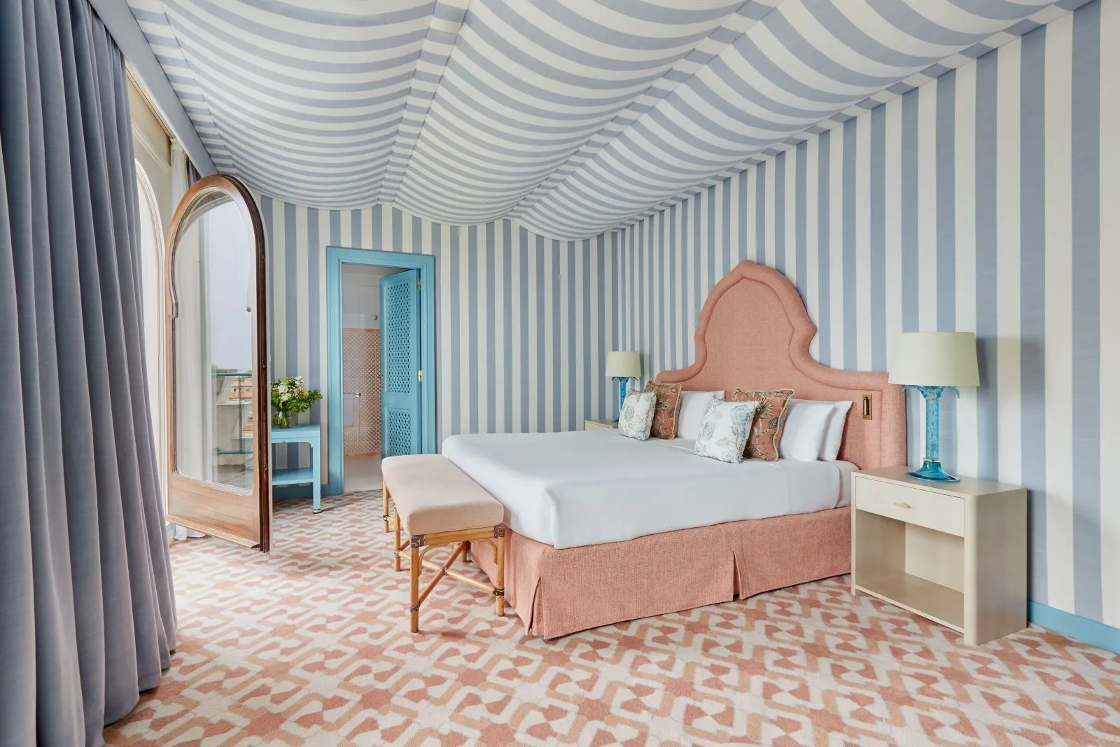 A blue-and-white striped room with a tented ceiling, a pink headboard and the arched door open onto the terrace.