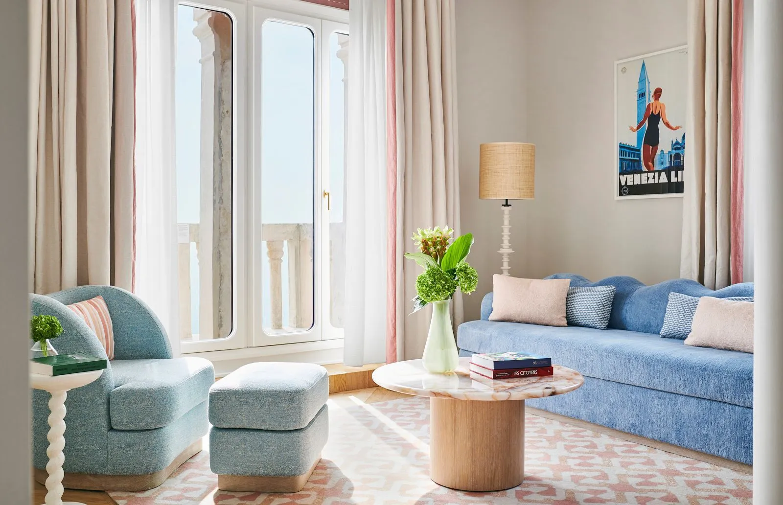 The suite’s sitting room: a blue sofa, a pale-blue armchair, a pink marble table, a vintage Lido poster and French windows onto the balcony.