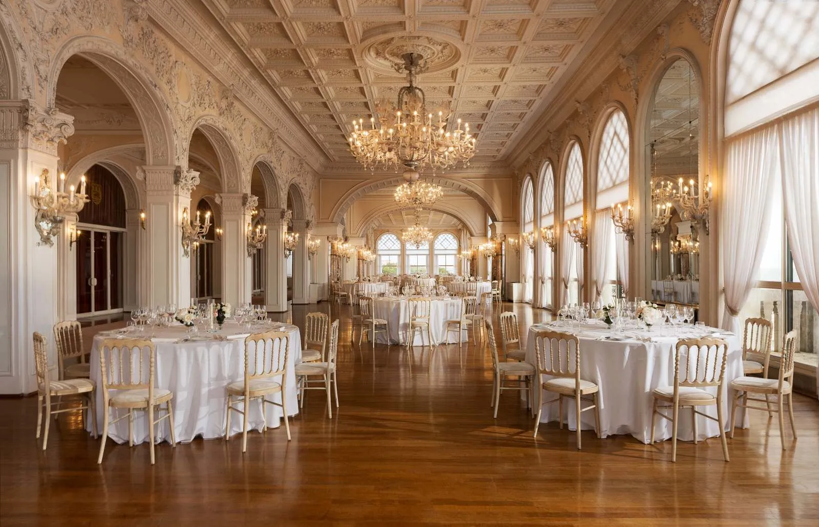 The Sala Stucchi laid for a banquet: round tables on parquet, glass chandeliers, a coffered ceiling and arched windows onto the sea.