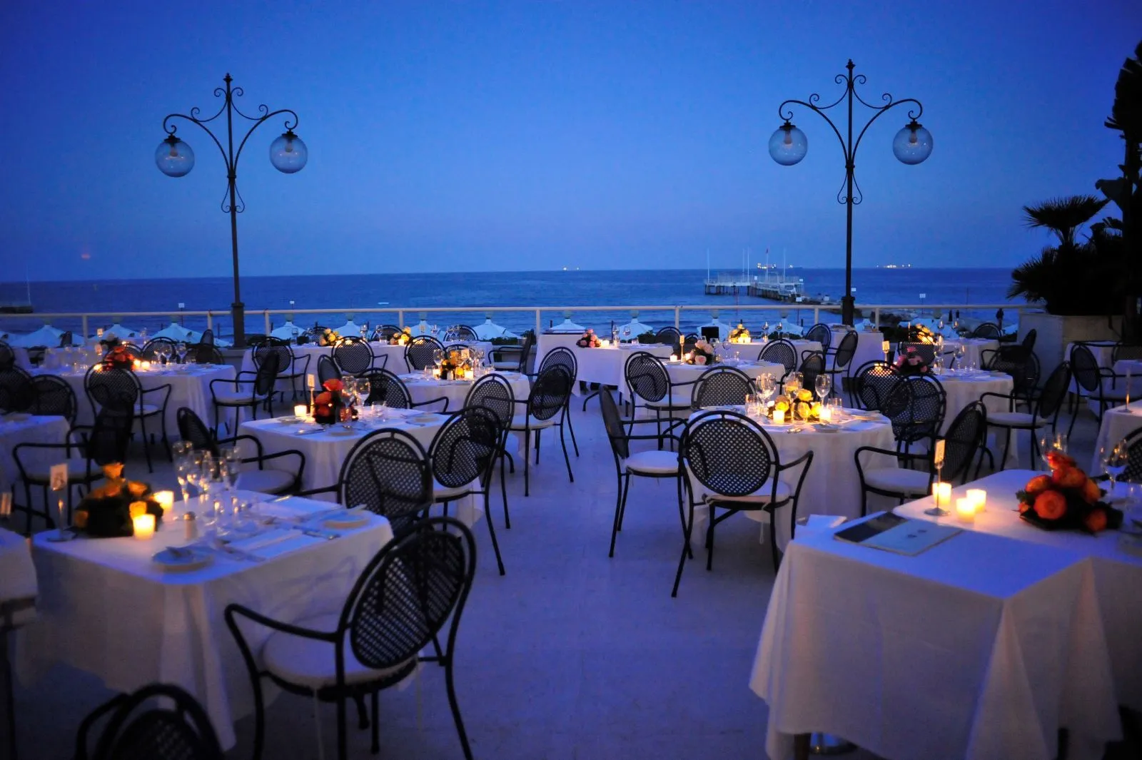 The terrace at dusk: tables with candles and flowers, globe lamps and the blue sea beyond the balustrade.
