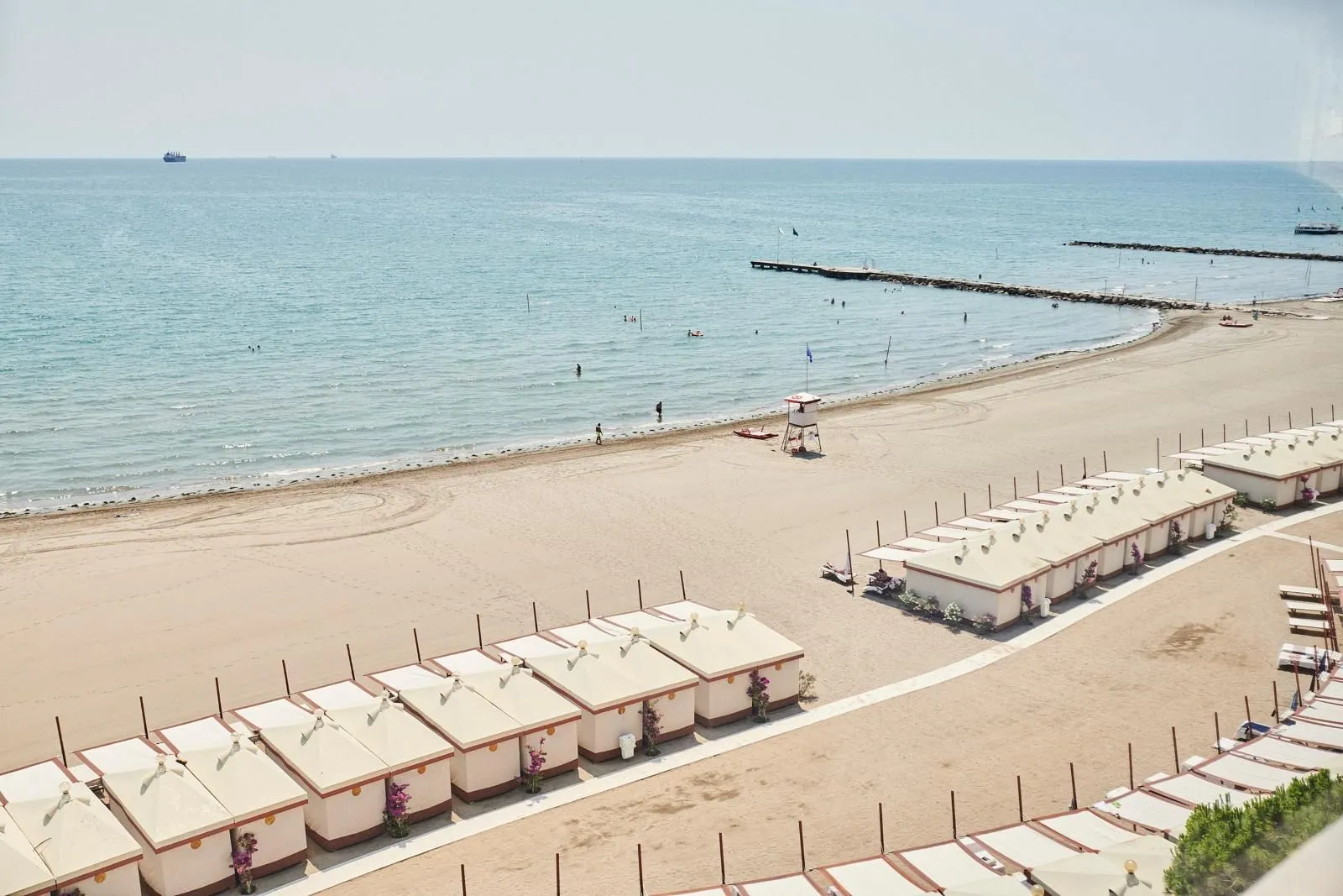 The beach from above: 2 rows of white cabanas on the sand, the lifeguard tower and the calm sea out to the jetties.