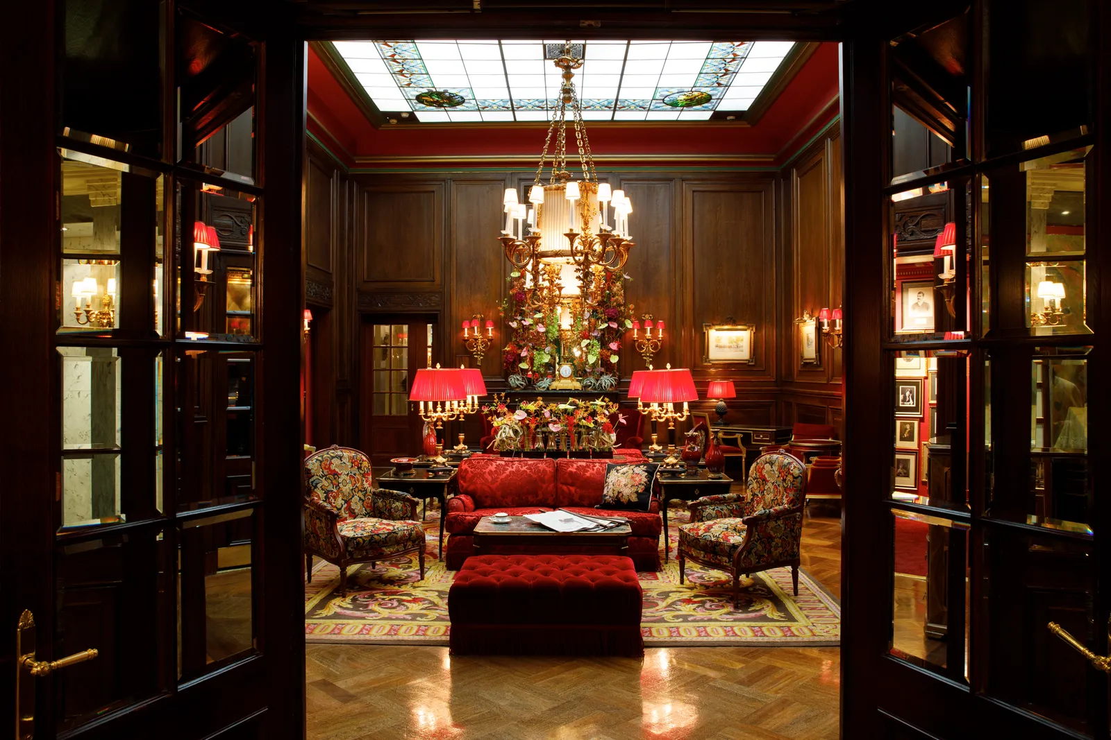 The lobby seen through open doors: dark panelling, red sofas and lampshades, a chandelier and the glass skylight.