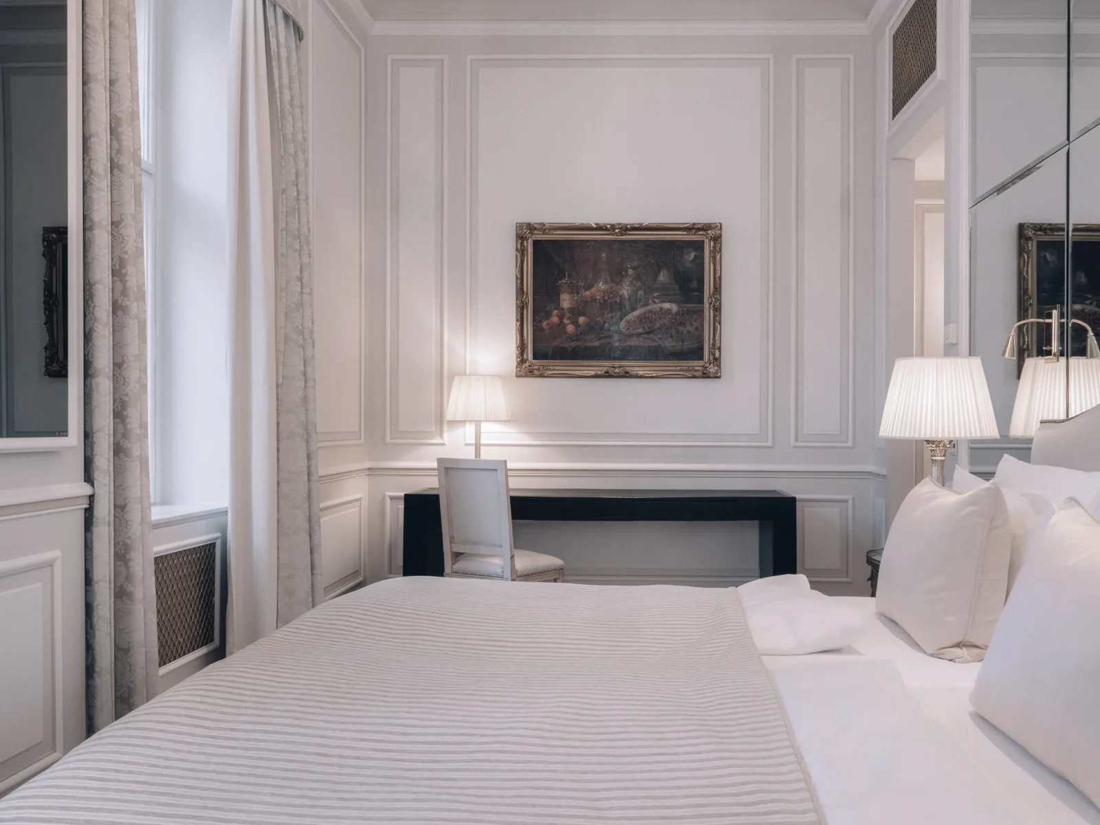 A Deluxe Room: the white bed, pale panelled walls, a painting above the desk and striped curtains at the window.
