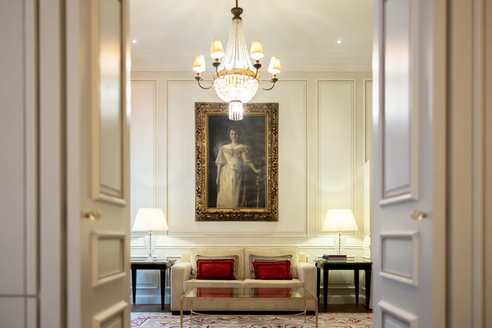 A Family Suite sitting room: a pale sofa with red cushions beneath a large gilt-framed portrait and a crystal chandelier.