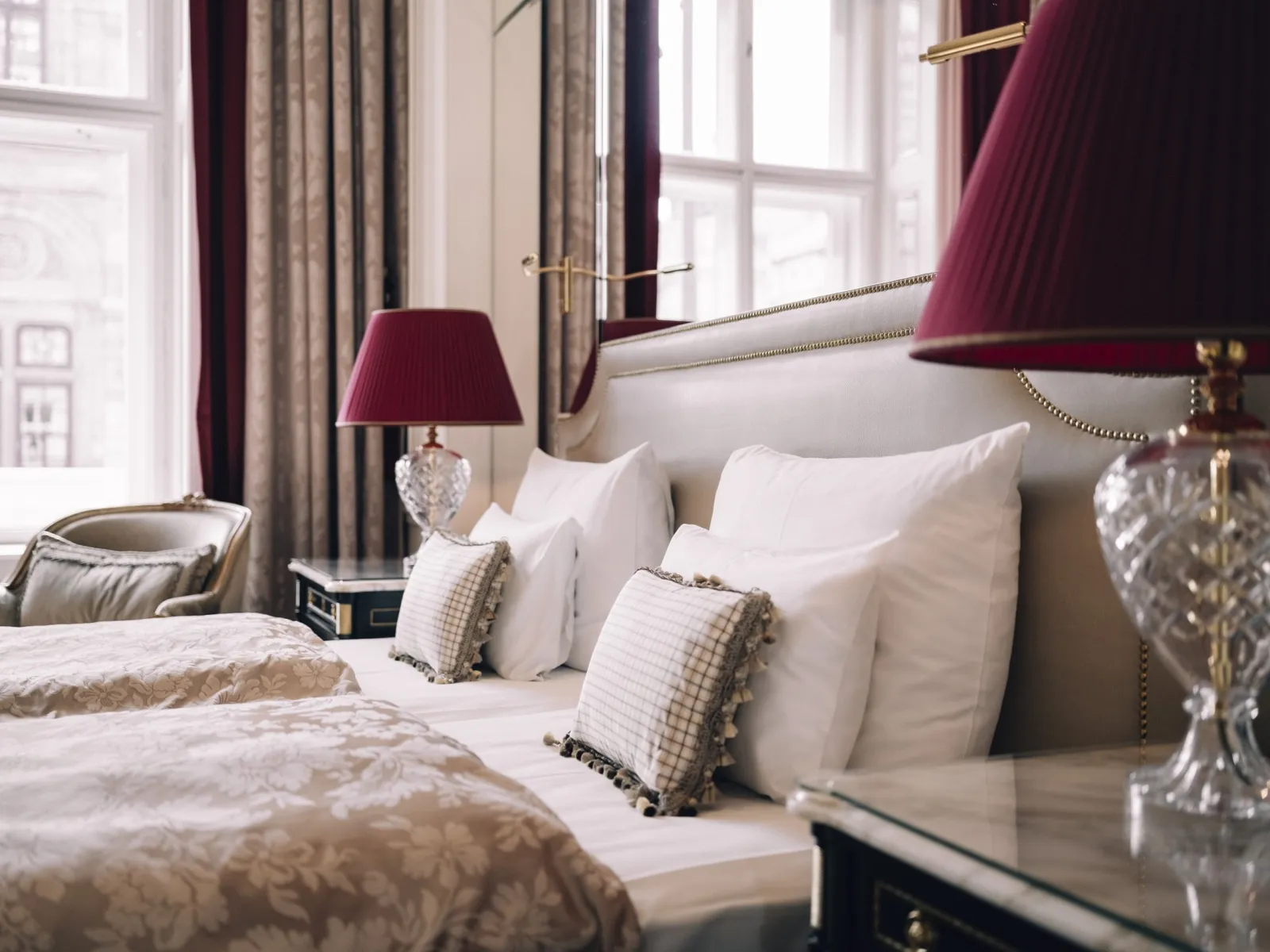 The bedroom of a Grand Signature Suite: twin beds side by side with pale cushions, crystal lamps with red shades and 2 tall windows.