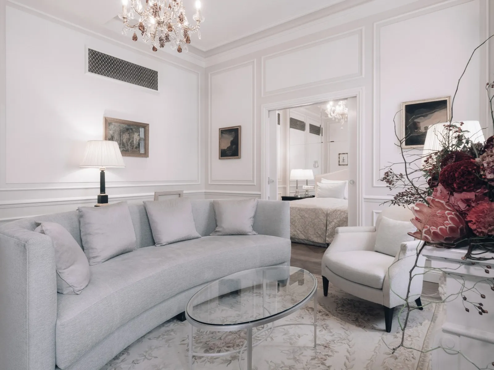 A Junior Suite sitting area: a pale curved sofa, a glass table, white panelled walls and the bedroom through the doorway.