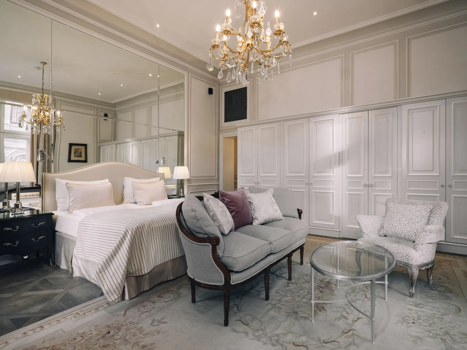 A Signature Suite: the bed with a pale headboard, a small sofa at its foot, a wall of white wardrobes and the crystal chandelier.