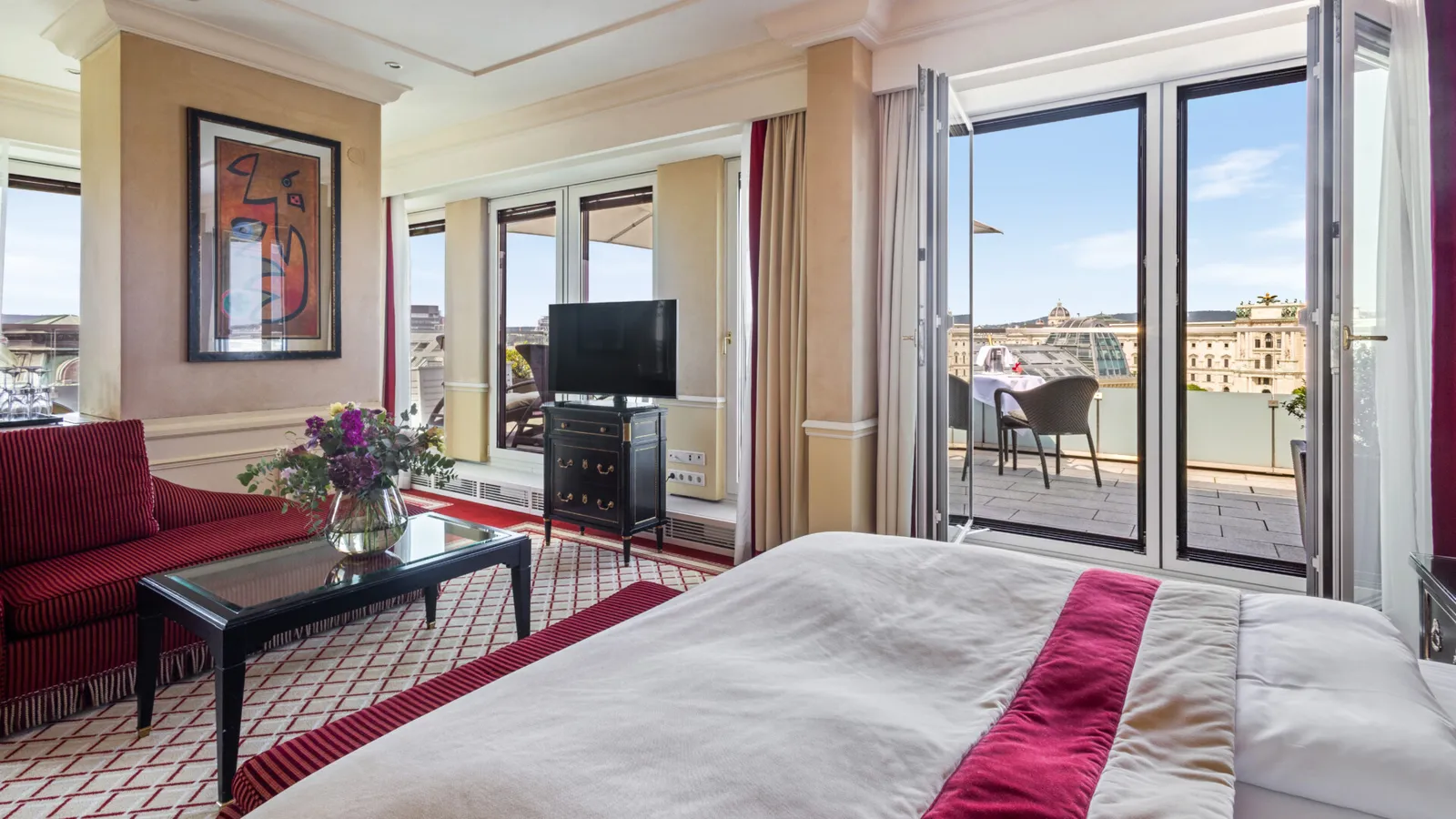 A Signature Suite with Terrace: the bed with a red runner, a red sofa and French windows open on the terrace above the rooftops.