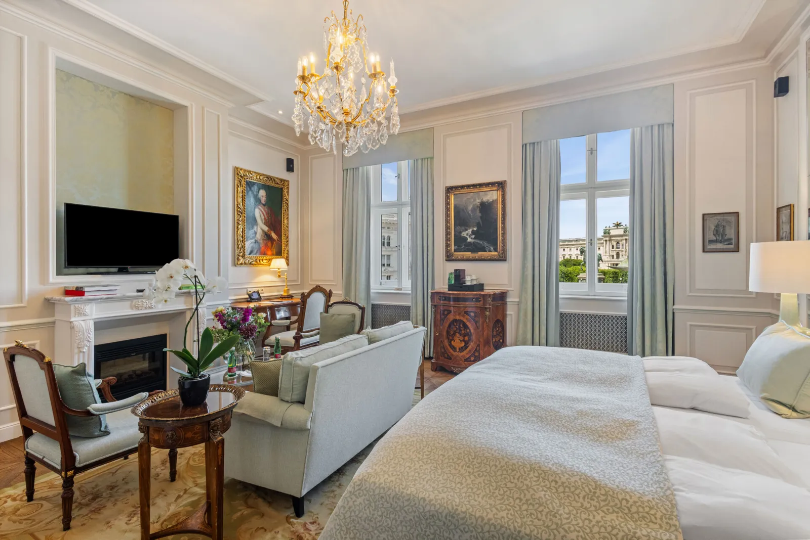A Signature Suite with view: the bed, a sofa and an armchair by the fireplace, pale-blue curtains and 2 windows on the city.