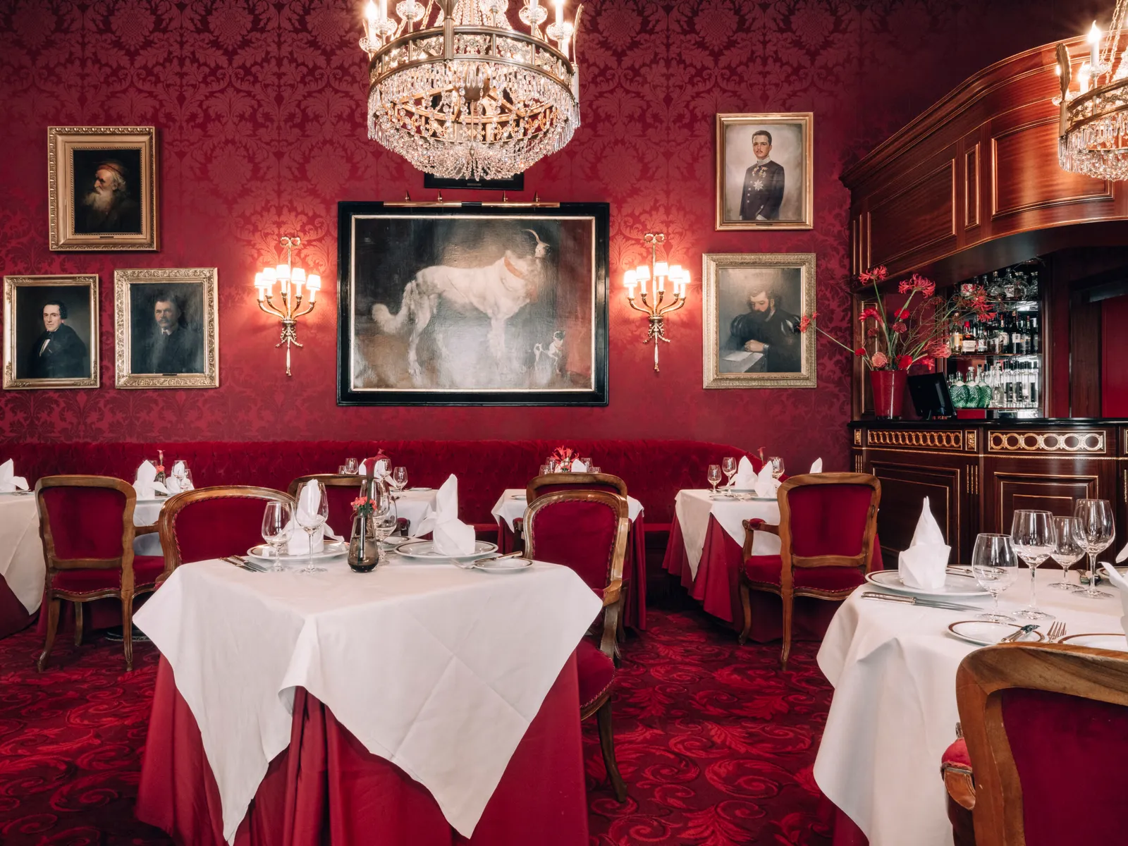 The Rote Bar dining room: red damask walls, oil portraits, velvet chairs and white-clothed tables under a crystal chandelier.