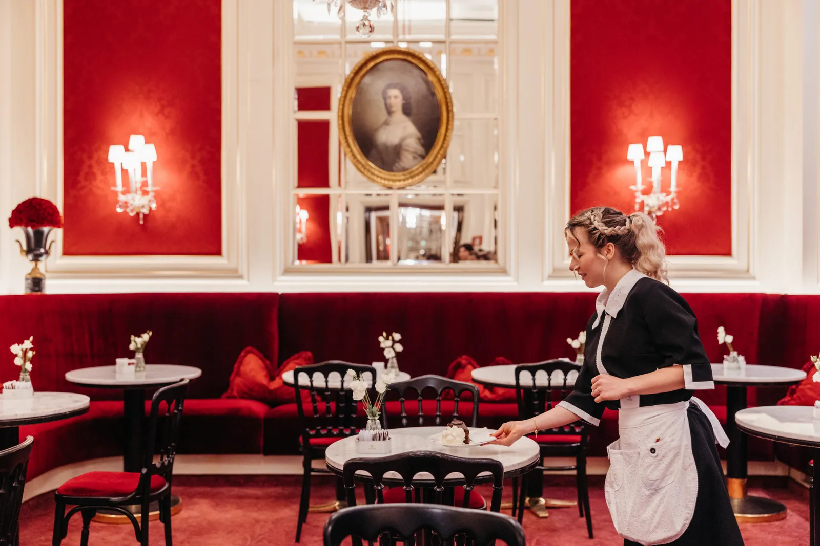 A waitress in a white apron sets a slice of Original Sacher-Torte on a marble table at Café Sacher, before the red velvet banquette.