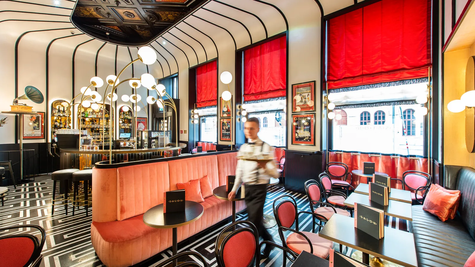 Salon Sacher: a coral banquette curving round the bar, globe lamps, red blinds at the windows and a black-and-white patterned floor.