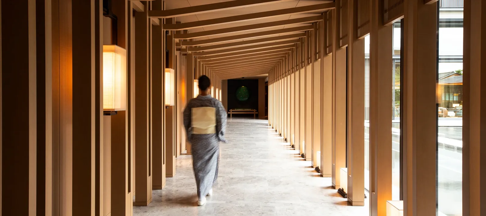 A woman in kimono walks the long timber corridor, between slender pillars and wall lanterns.