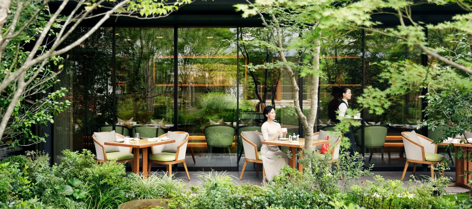 FORNI’s terrace in the garden: tables and armchairs among the trees and ferns, with 2 guests seated before the glass walls.