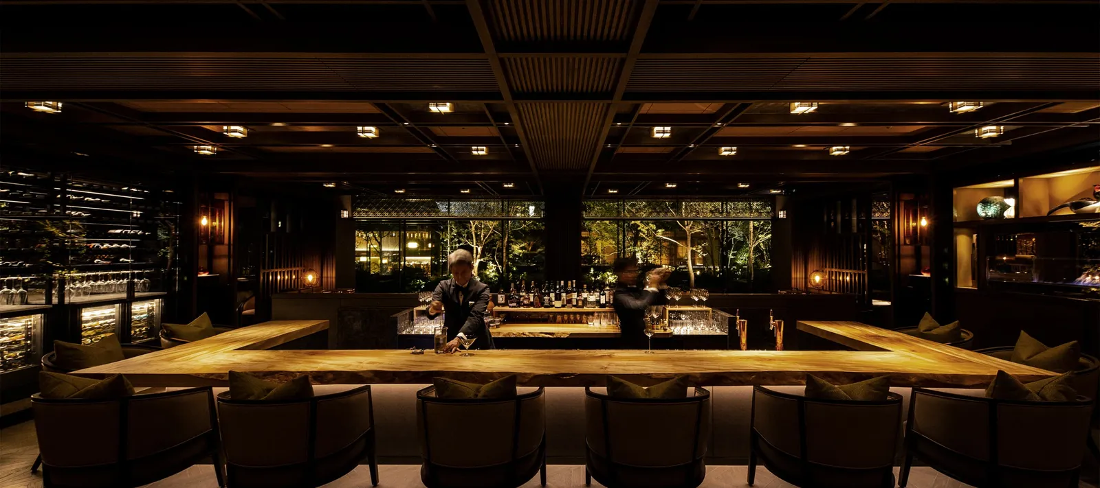 THE GARDEN BAR at night: the long wooden counter, the lit shelves of bottles and the glass walls onto the dark garden.