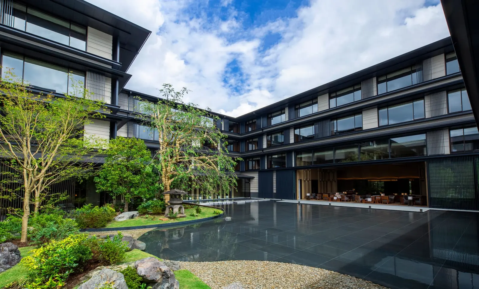 The garden from the edge of the pond: the rocks, the young trees and the hotel’s 4 wings under a blue sky.