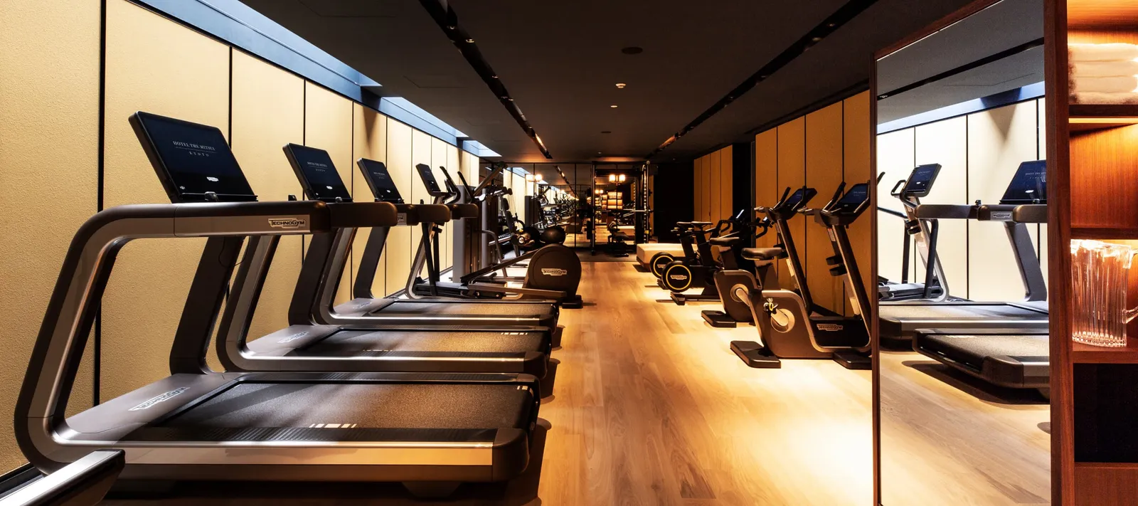 The gym: a row of treadmills and bikes on the wooden floor, under a dark ceiling and warm lights.