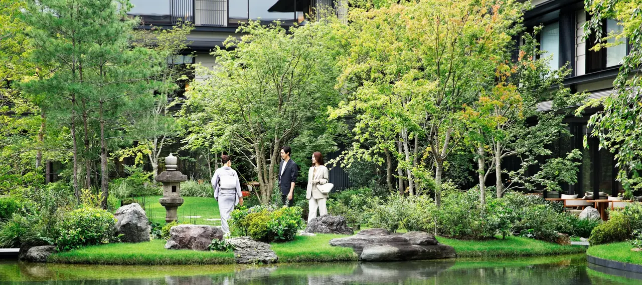 2 guests in pale clothes walk among the rocks, stone lanterns and maples of the garden, beside the pond.