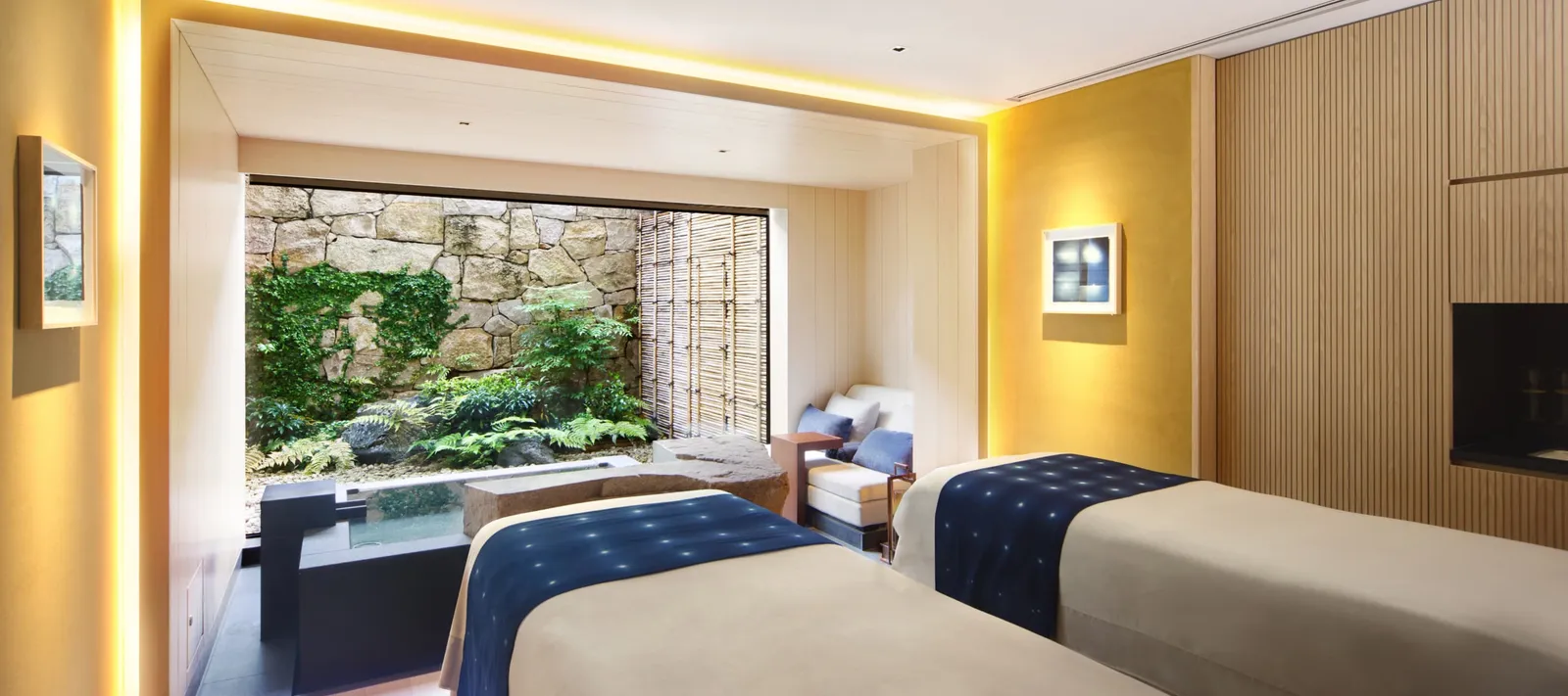 A treatment room with 2 beds and a wide window onto a small garden of stone and green.