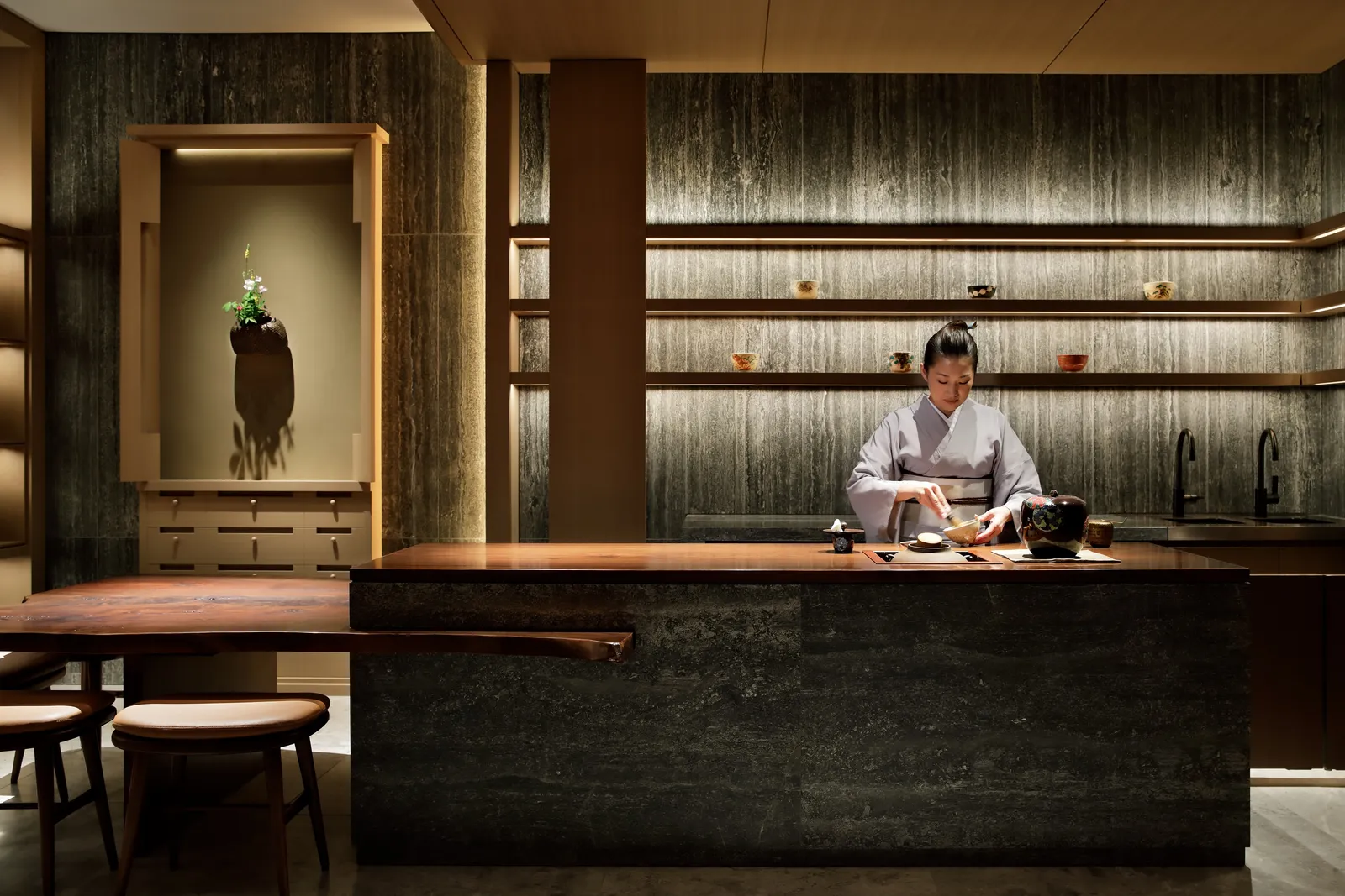 The tea room: a woman in kimono whisks matcha at the stone counter, before lit wooden shelves.