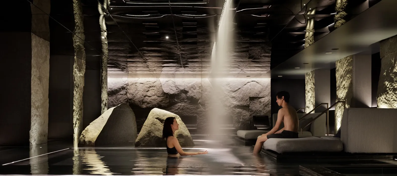 The underground thermal spring: a waterfall drops between 2 boulders into a dark stone pool, where 2 guests in swimwear relax.